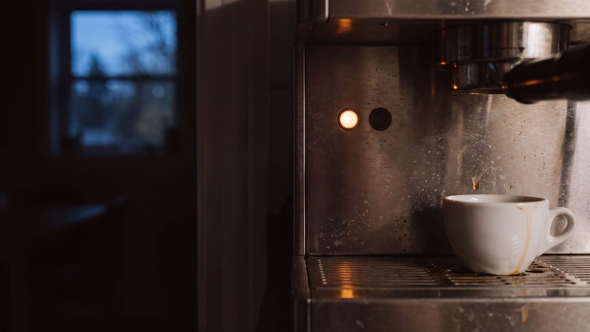 A single amber indicator light glowing beside a dark one on a stainless espresso machine in a dark kitchen, a stained cup below